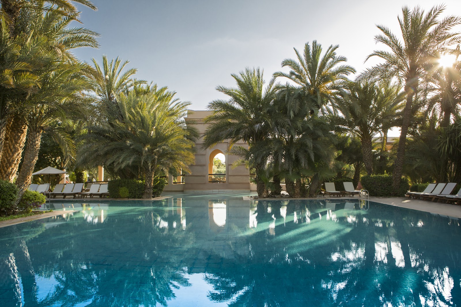 Large resort pool surrounded by tall palm trees, with lounge chairs and a beige building with arched entrance in the background.