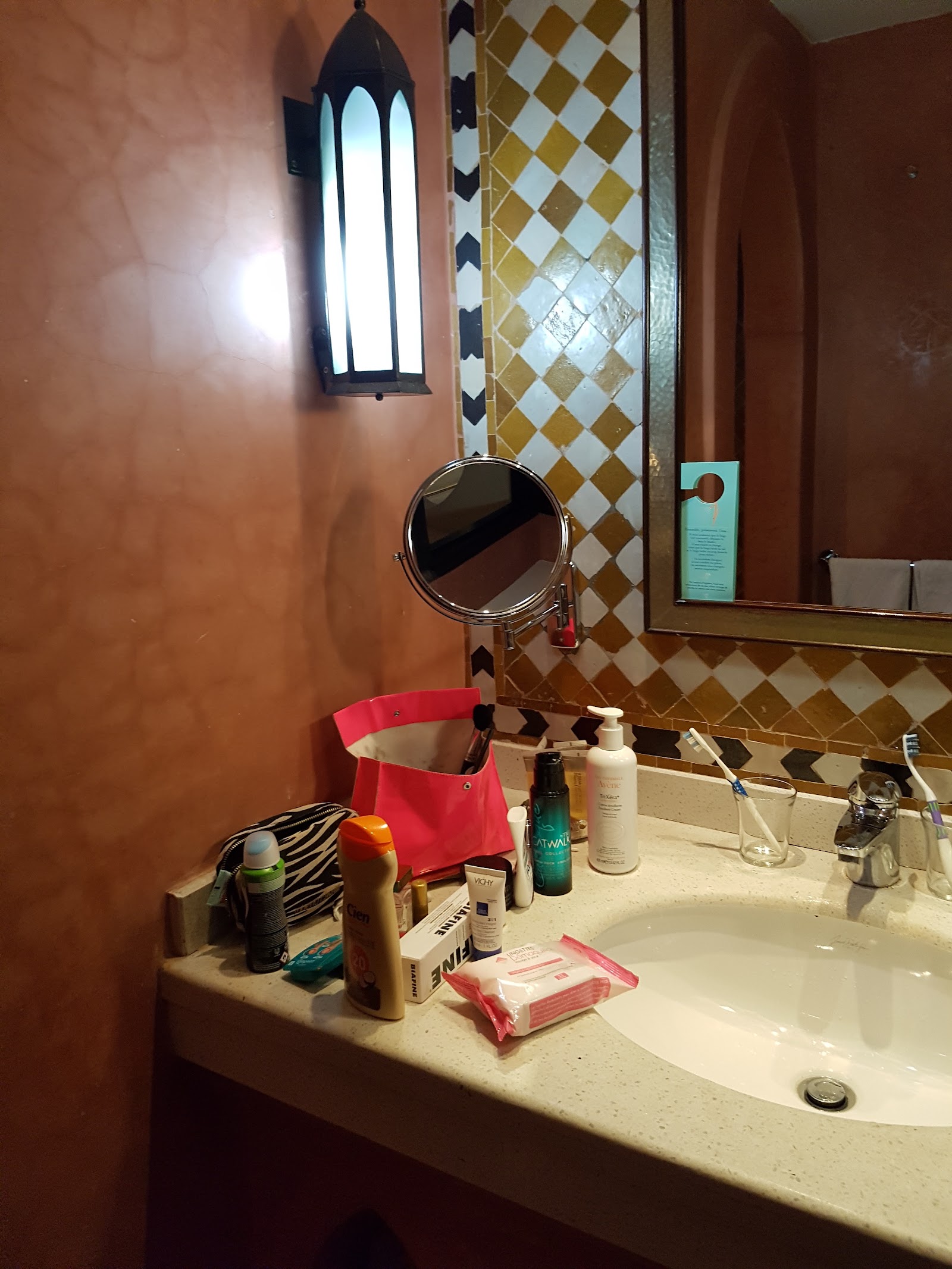 Bathroom vanity with white sink, round mirror, Moroccan-style sconce light, terracotta walls, and gold and white diamond-patterned tile backsplash with toiletries arranged on counter.