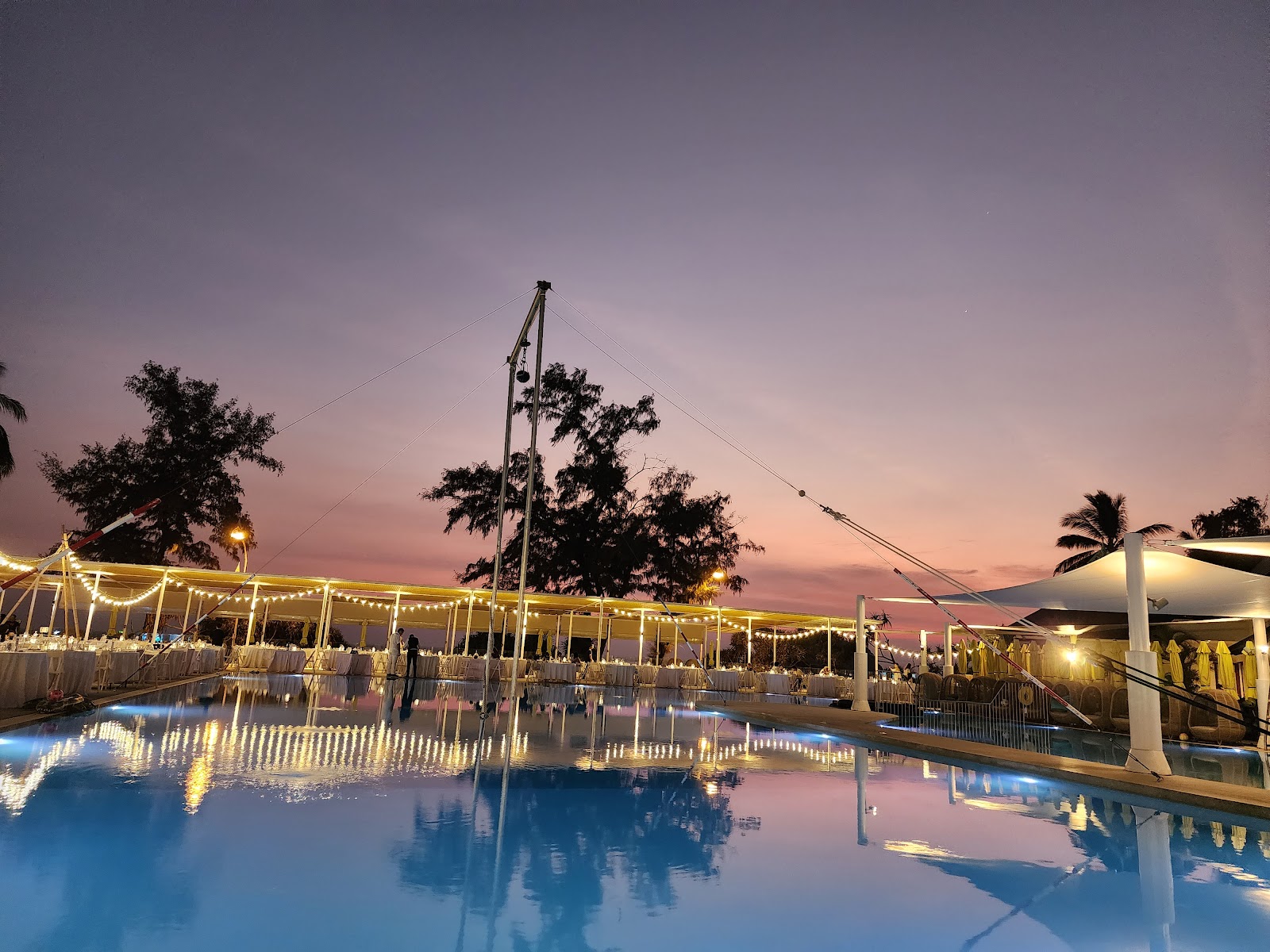 Illuminated pool deck at dusk with string lights strung across a white tent structure, palm trees silhouetted against a pink and purple sky.