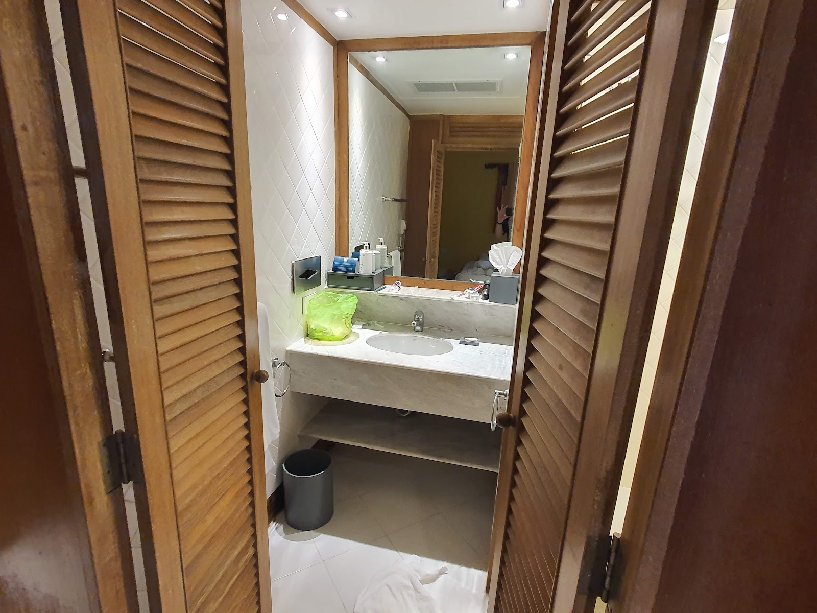 Resort bathroom with wooden louvered doors, white tile walls, countertop sink with mirror, green waste bin, and toiletries arranged on the counter.