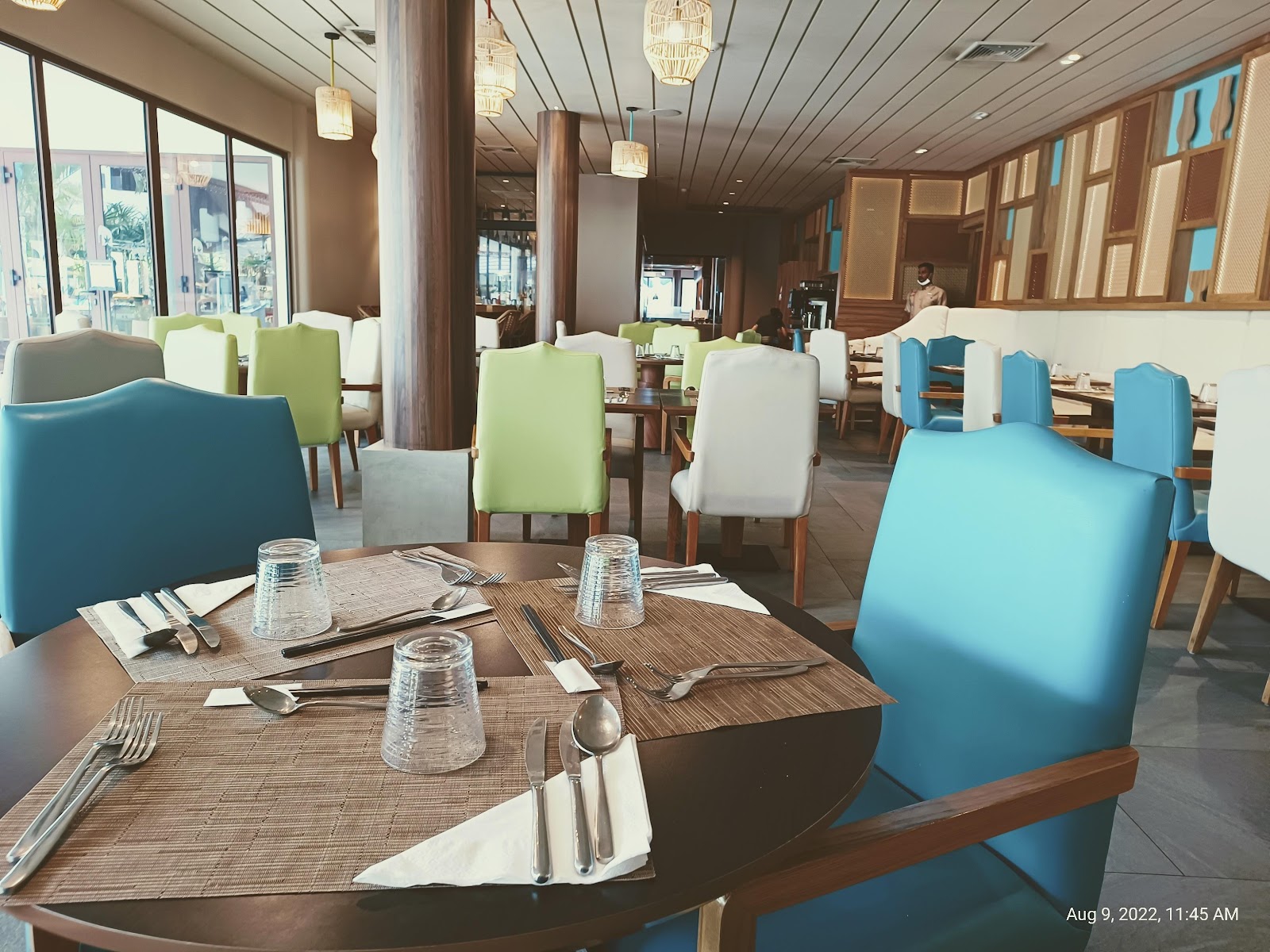 Modern dining room with turquoise and green chairs at tables set with place settings, water glasses, and napkins under pendant lights and large windows.