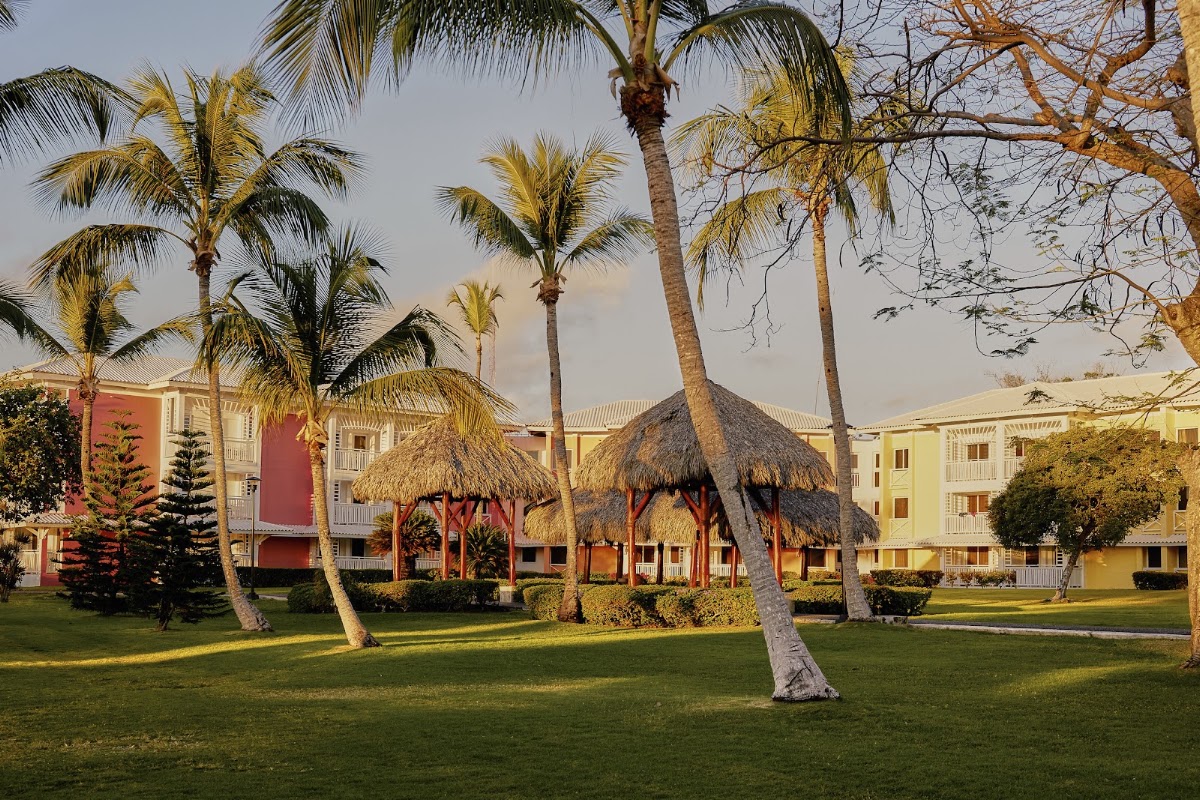 Palm trees shade the green lawn in front of colorful resort buildings with thatched-roof structures at Club Med Punta Cana.