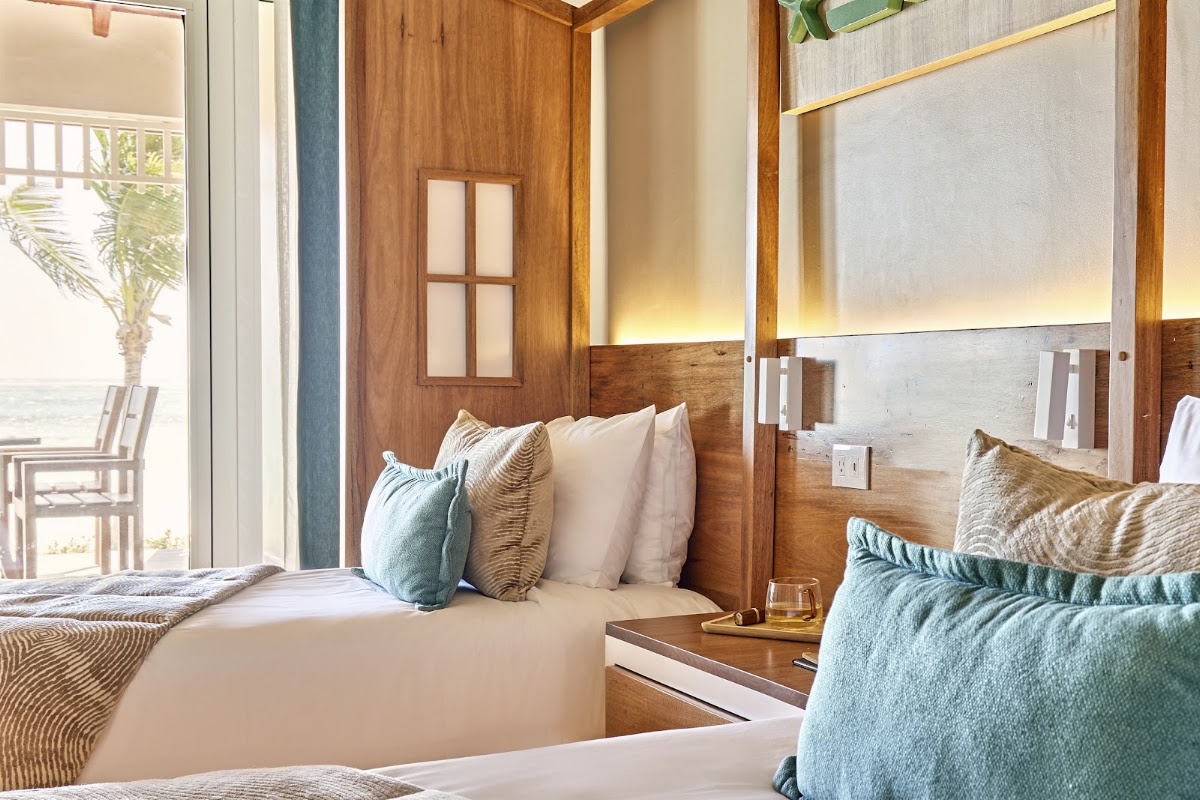 Resort guest room with two beds, wooden accents, ocean view through glass doors, and palm tree visible on the balcony.