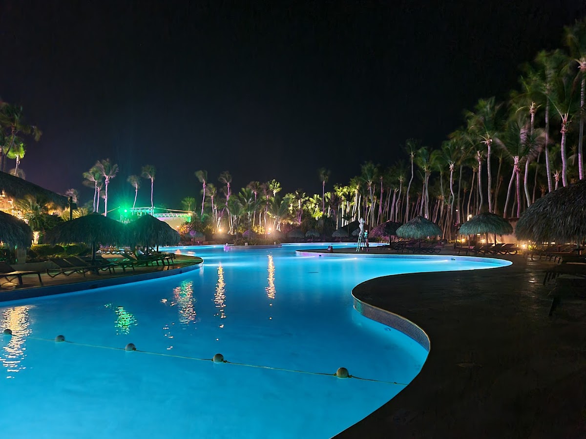 Illuminated resort pool glowing blue at night, lined with palm trees, thatched cabanas, and lounge chairs at Club Med Punta Cana.