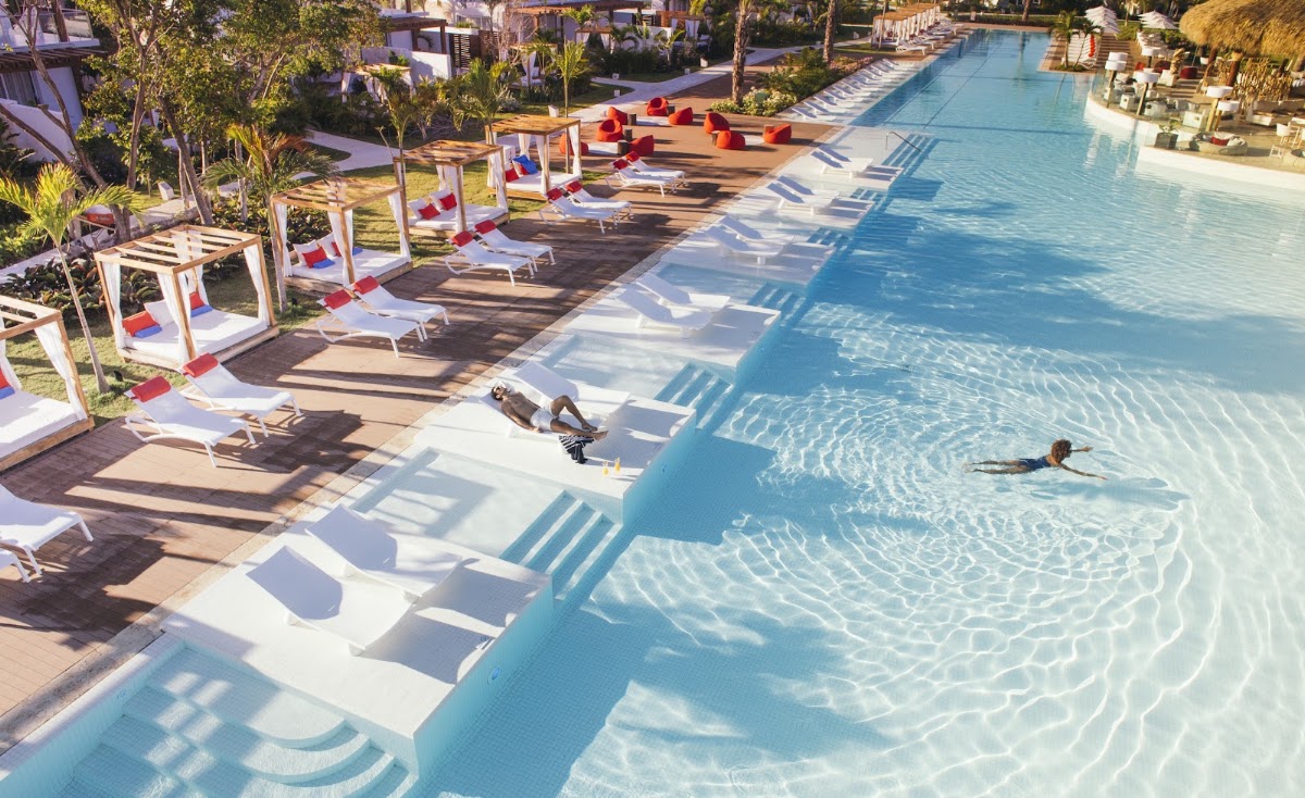 Guests swim and lounge on white sunbeds beside a clear blue pool at Club Med Punta Cana, with red-accented cabanas and tropical palms lining the deck.