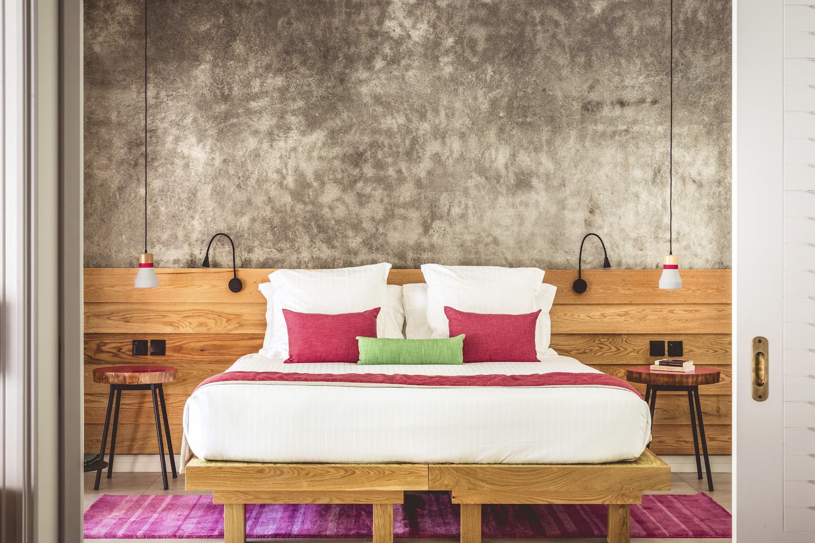 Modern resort bedroom with king bed, red and green pillows, wooden headboard with pendant lights, and purple area rugs on wood flooring.