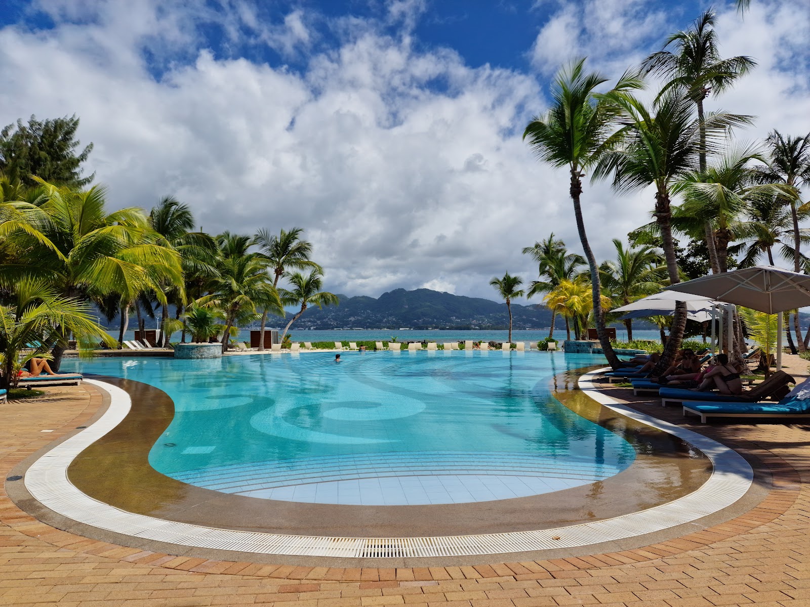 Resort pool with curved edges and turquoise water, surrounded by palm trees, lounge chairs, and umbrellas, with mountain and ocean views beyond.