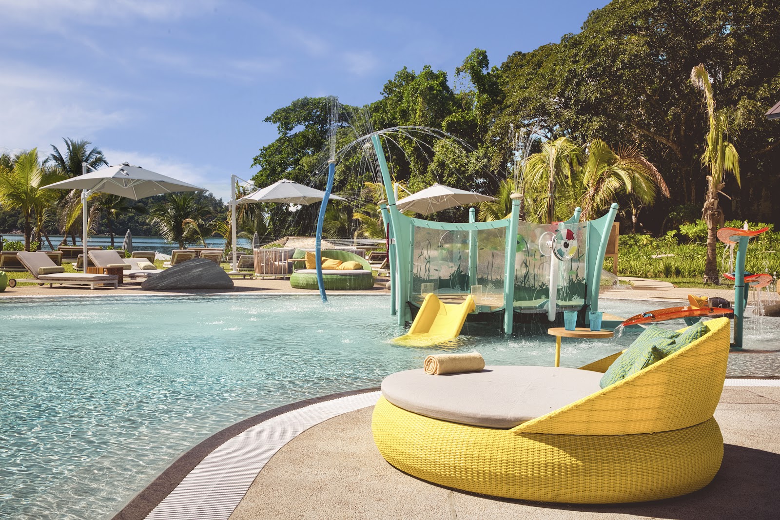 Yellow lounge chair beside a shallow pool with children's water play structure and palm trees at Club Med Seychelles.