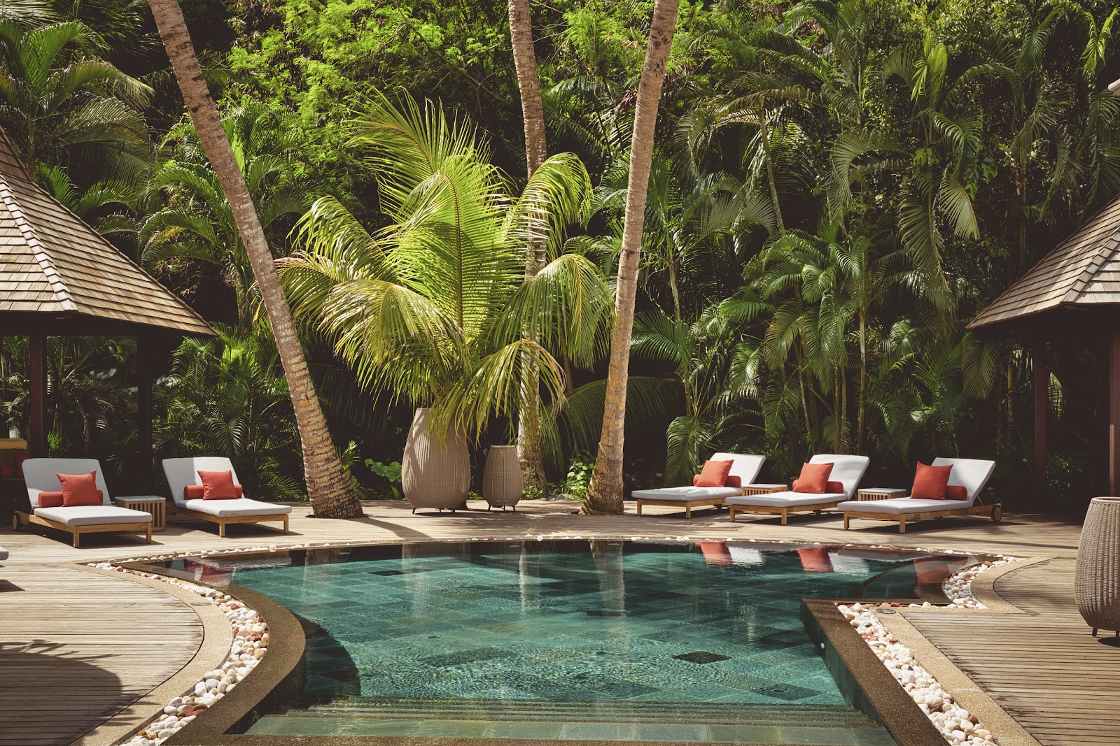 Small pool surrounded by wooden loungers with coral cushions, tall palm trees, and thatched-roof bungalows at Club Med Seychelles.