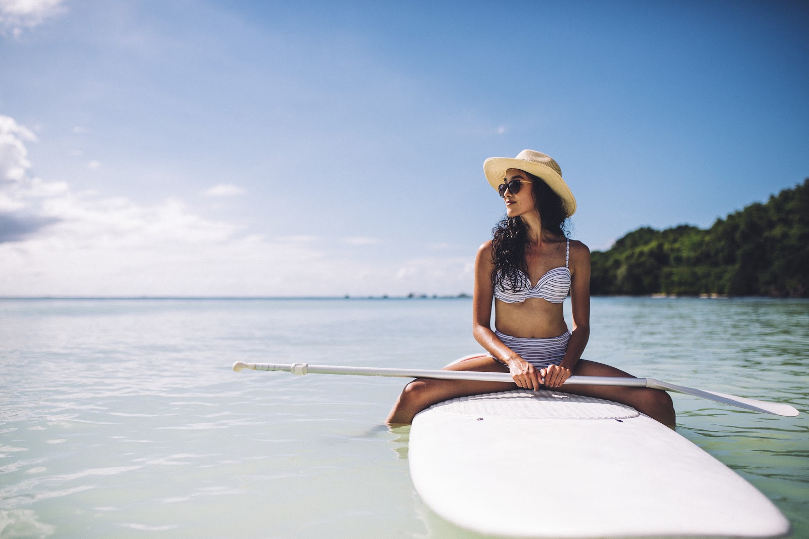 Woman in striped bikini and straw hat sits on paddleboard in shallow turquoise water with forested shoreline behind.