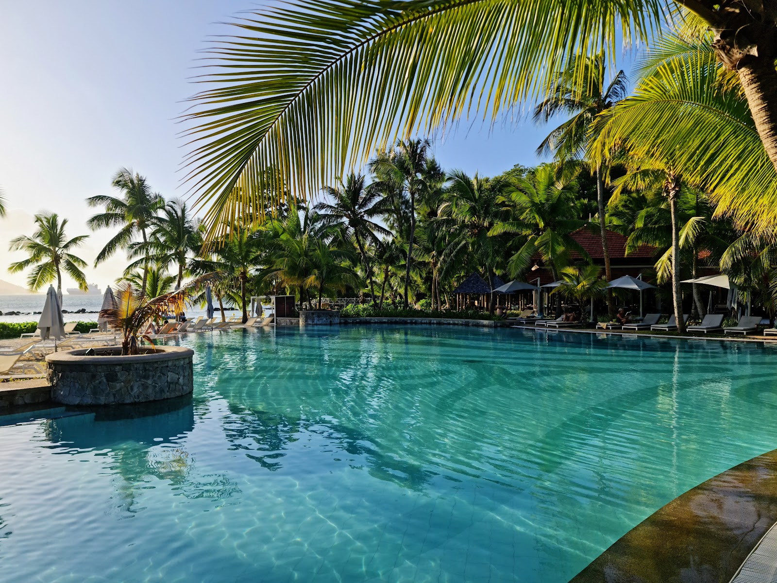Turquoise pool surrounded by palm trees with loungers and umbrellas at Club Med Seychelles, ocean visible in background.