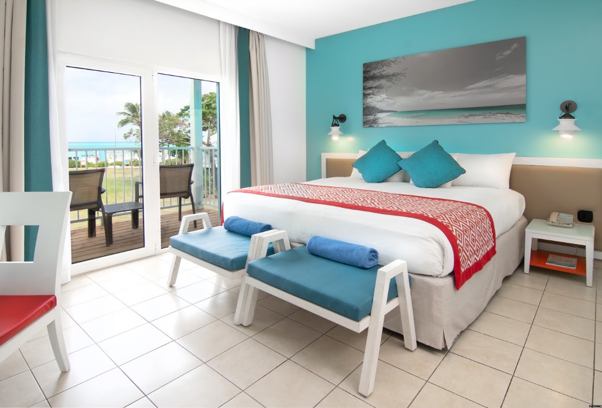 Beachfront bedroom at Club Med Turkoise with turquoise walls, king bed with red runner, balcony overlooking ocean and palm trees.