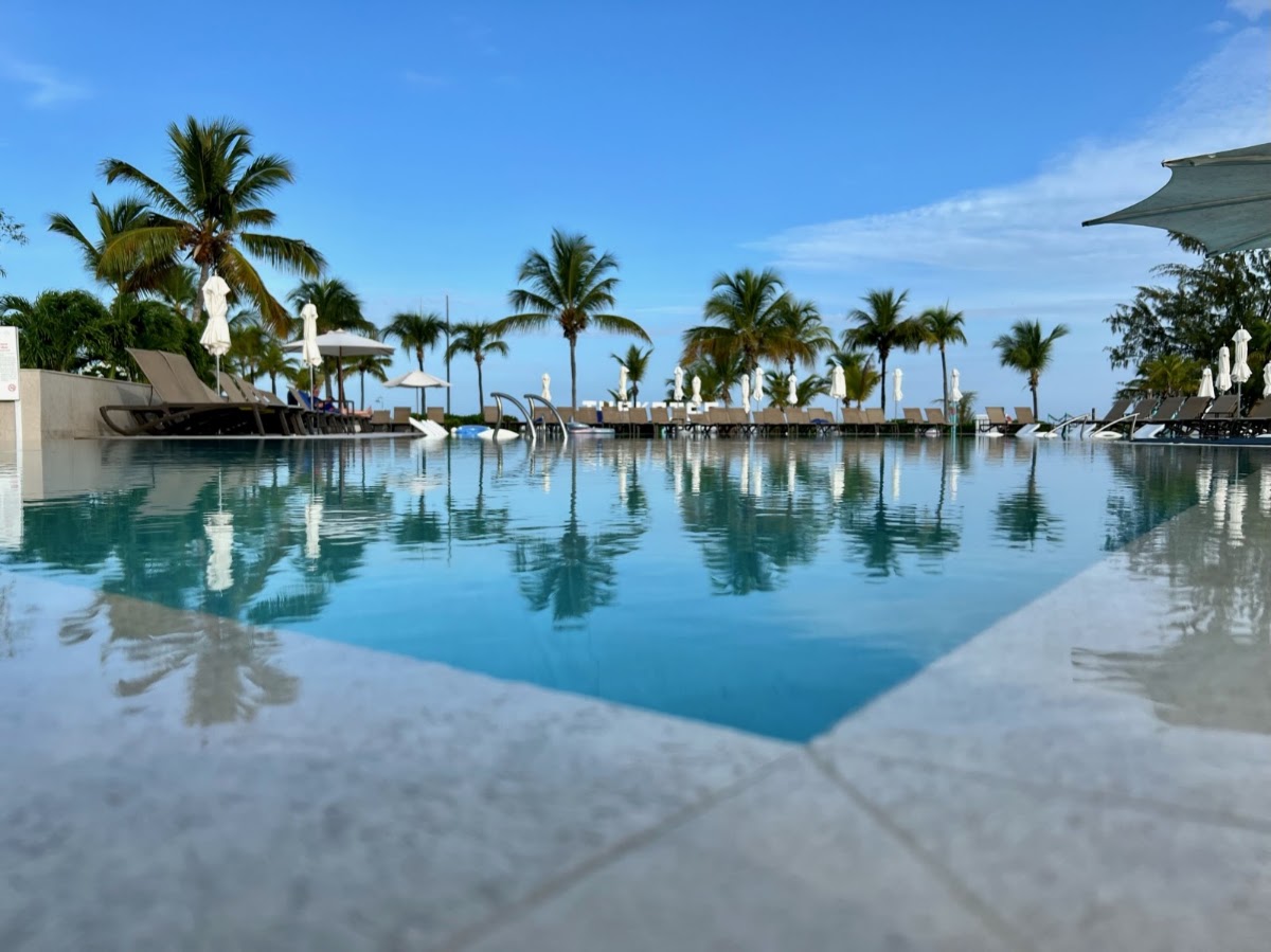 Resort pool with pale stone deck, lounge chairs, palm trees, and calm turquoise water under clear sky at Club Med Turkoise.