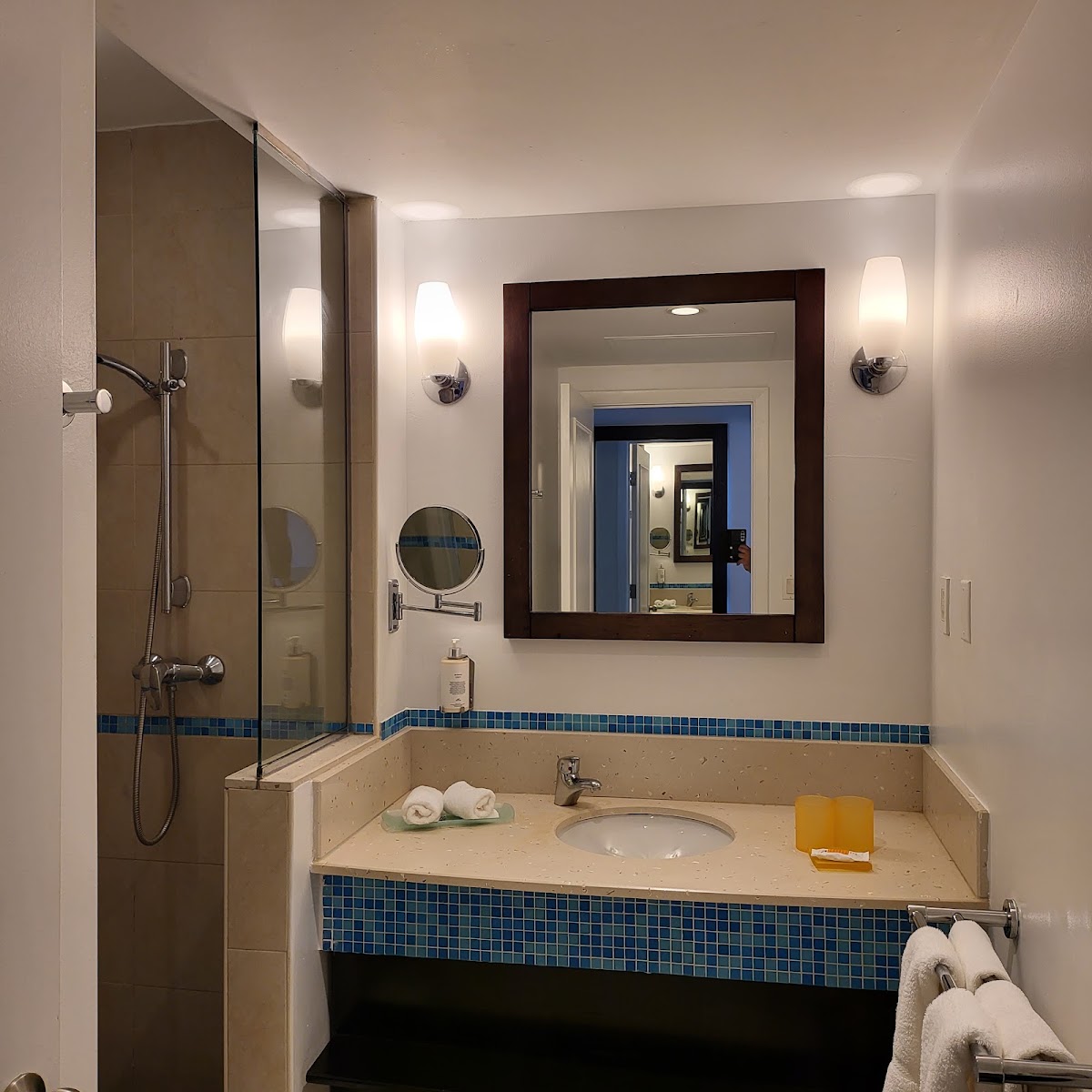 Resort bathroom with walk-in shower, framed mirror, beige countertop with blue tile accents, and rolled towels.