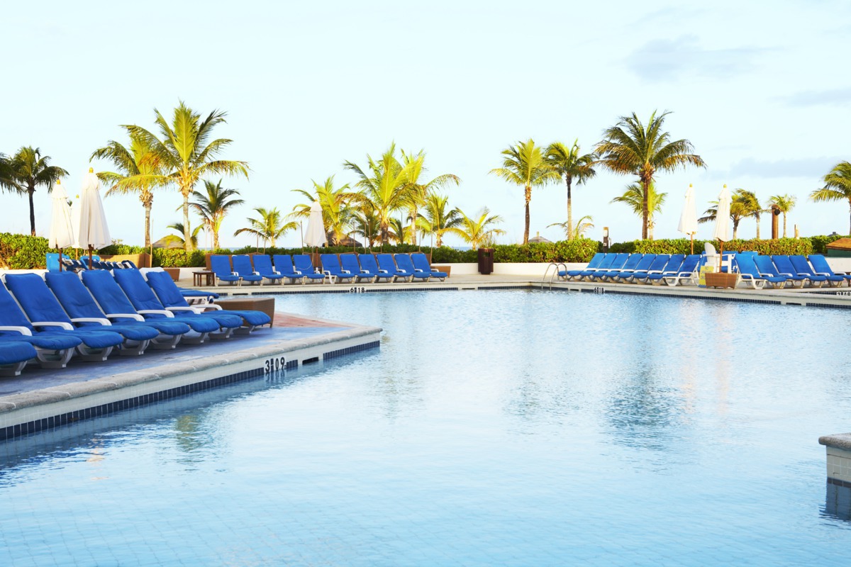 Club Med Turquoise pool with rows of blue lounge chairs, palm trees, and white umbrellas under clear sky.