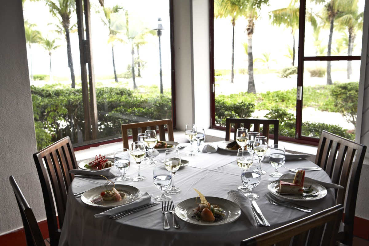 Dining table set for multiple guests with plated entrees, wine glasses, and water glasses, overlooking palm trees and manicured grounds through large windows at Club Med Turkoise.