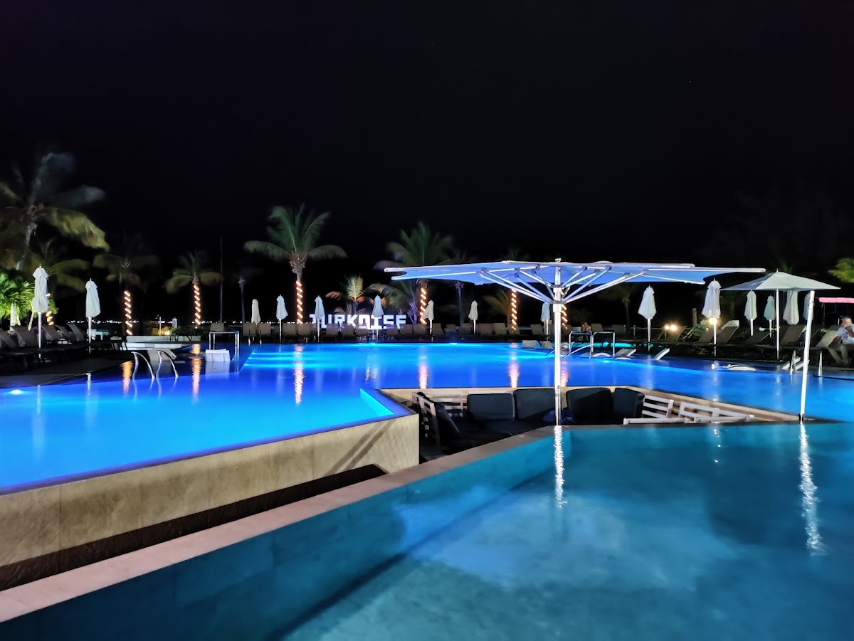 Club Med Turkoise pool at night with blue-lit water, white umbrellas, palm trees, and lounge chairs under a dark sky.
