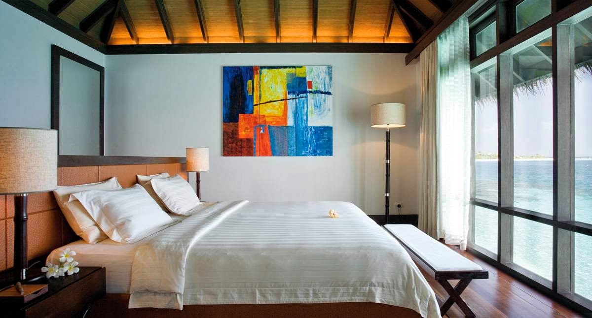 Modern bedroom at Coco Bodu Hithi with ocean views through floor-to-ceiling windows, tan leather headboard, and colorful abstract artwork.