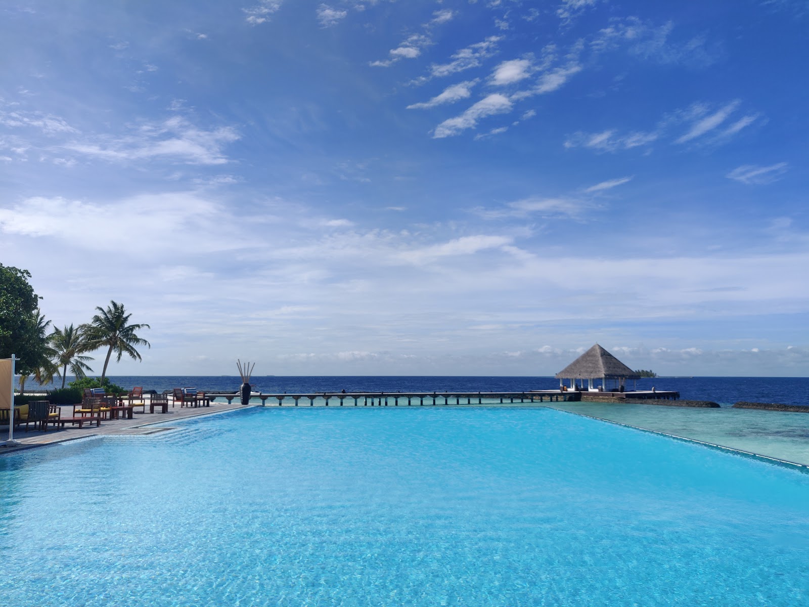 Turquoise pool and ocean at Coco Bodu Hithi with a thatched-roof overwater bungalow and wooden dock extending toward the horizon.