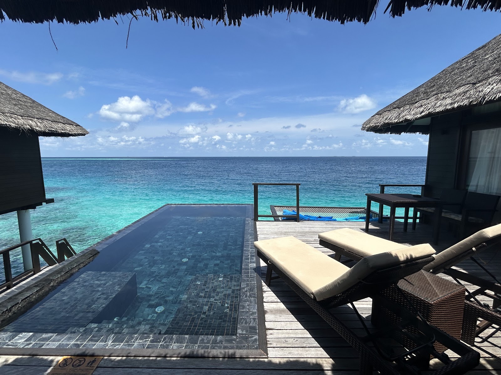 Overwater bungalows with thatched roofs flank an infinity pool overlooking turquoise lagoon and blue ocean at Coco Bodu Hithi.