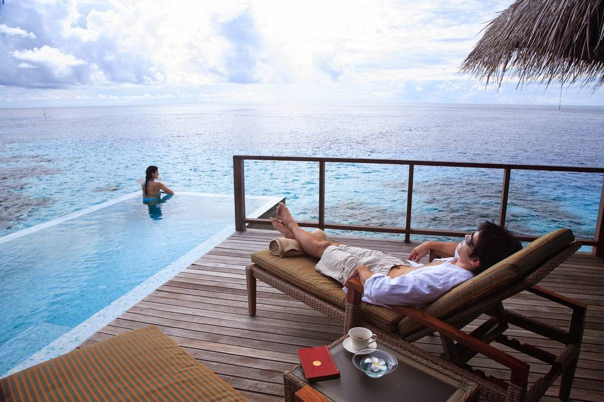 White lounge beds with yellow pillows line a wooden deck beside a turquoise pool overlooking the lagoon at Coco Bodu Hithi.