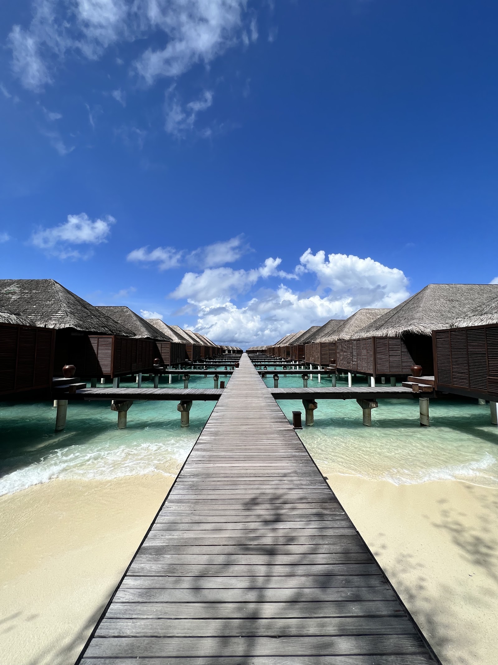 Aerial view of Coco Bodu Hithi island resort with overwater bungalows, white sand beach, palm trees, and turquoise lagoon surrounded by deep blue ocean.