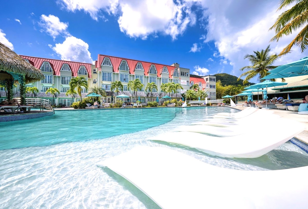 Turquoise pool and sandy beach front a colorful resort building with red roof and palm trees, under blue sky with white clouds.
