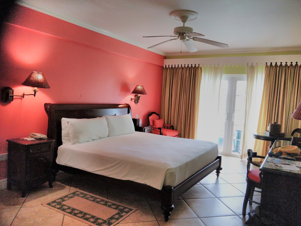 Resort bedroom with coral-pink accent wall, dark wood bed with white linens, ceiling fan, and glass doors opening to a balcony.