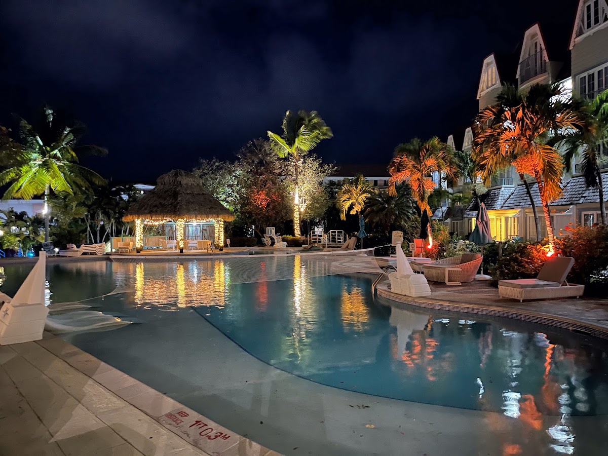 Nighttime pool area at Coco Palm Resort lit by warm uplighting on palms and thatched-roof bar, with resort buildings rising beyond.