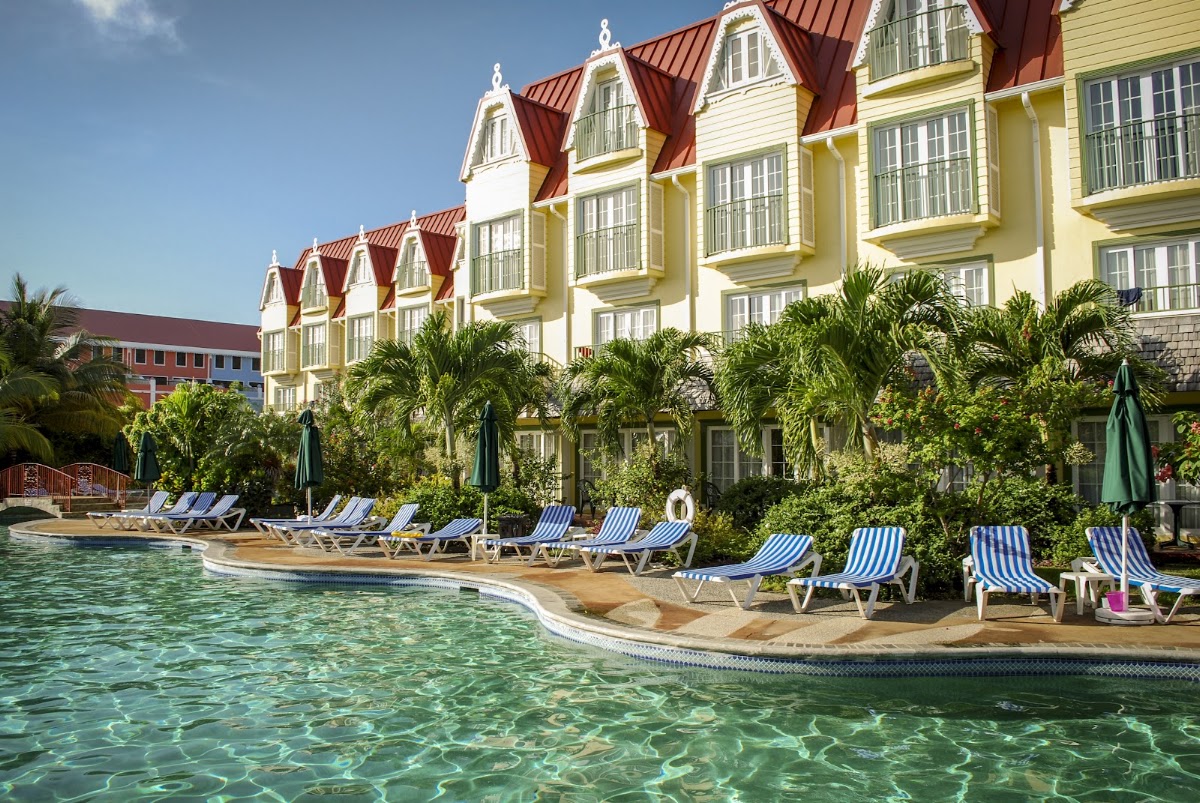 Yellow and white colonial-style buildings with red roofs overlook a pool with blue-striped loungers and tropical landscaping at Coco Palm Resort.