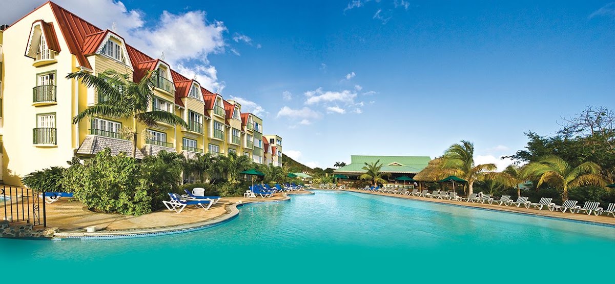 Yellow building with red metal roof beside turquoise pool lined with lounge chairs and palm trees under clear blue sky.