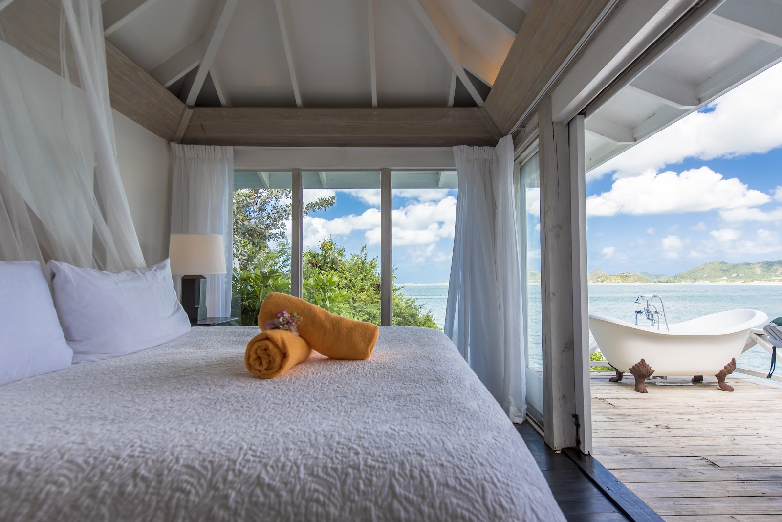 Beachfront villa bedroom at Cocobay Resort with four-poster bed, ocean views through glass doors, and soaking tub on the deck overlooking the sea.