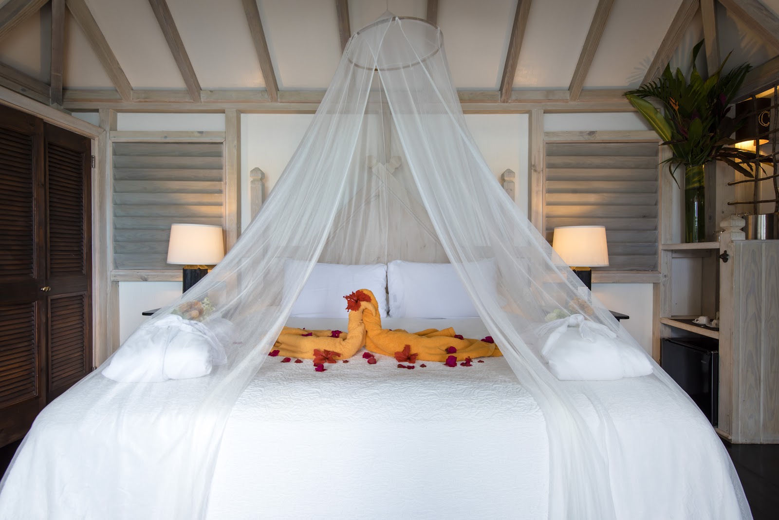 Canopied bed with white sheer curtains, yellow fabric décor, and scattered rose petals in a modern resort bedroom with louvered windows.