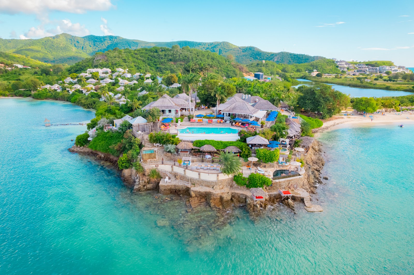 Aerial view of Cocobay Resort on a rocky peninsula surrounded by turquoise water, with a blue pool, thatched-roof buildings, and forested mountains beyond.