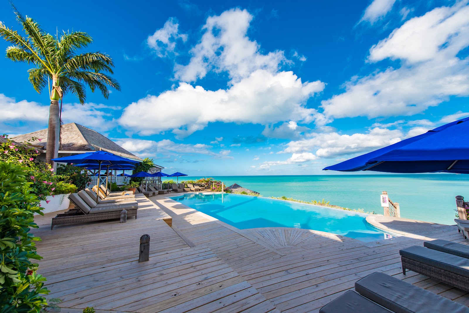 Wooden deck with lounge chairs and blue umbrellas overlooking an infinity pool that meets turquoise ocean at Cocobay Resort Antigua.