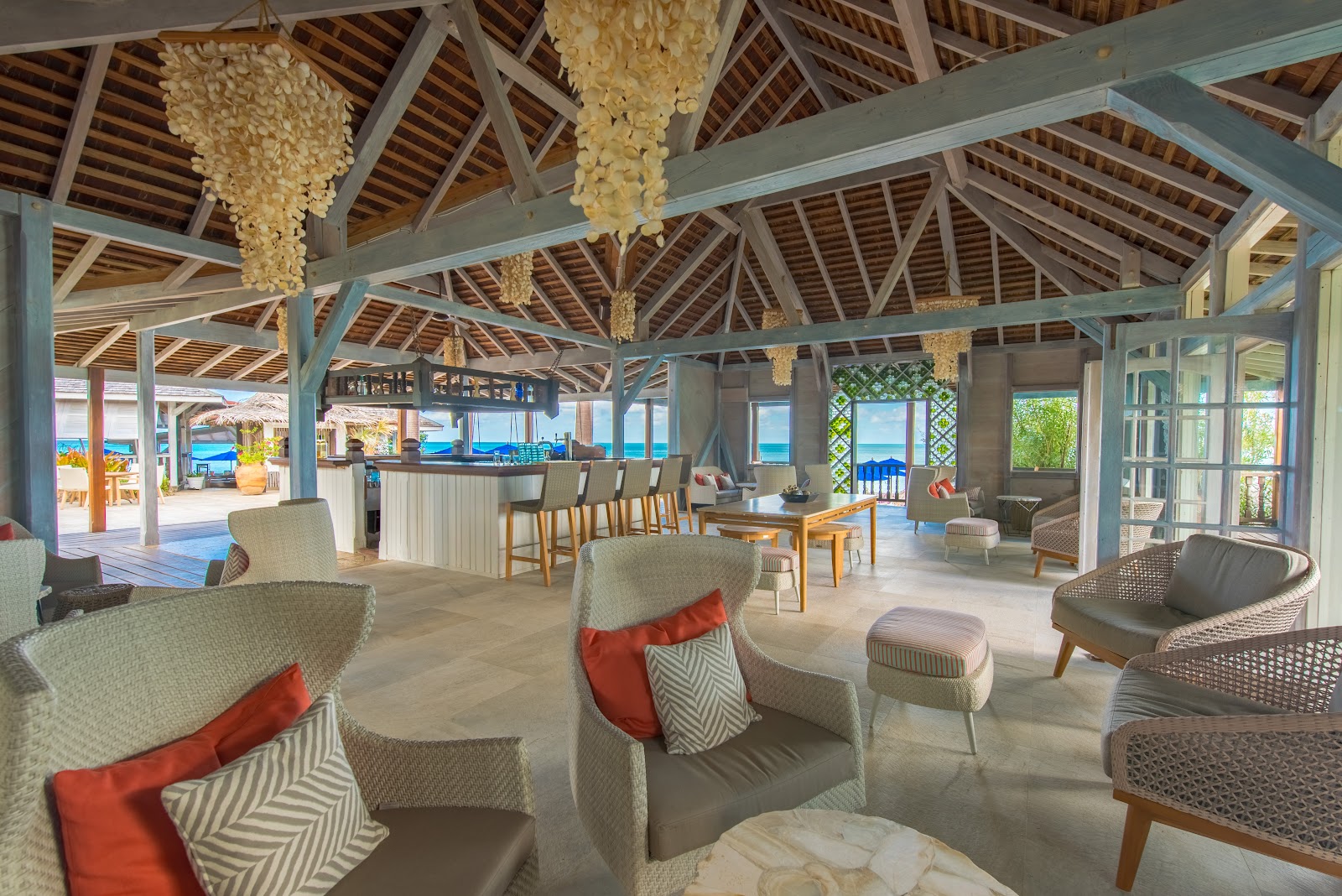 Open-air lounge with upholstered seating, coral accent pillows, hanging shell chandeliers, and ocean views through open doors at Cocobay Resort.