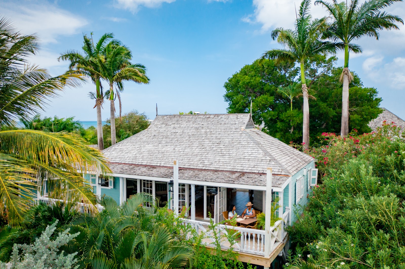 Light blue villa with gray pitched roof surrounded by tall palms and flowering shrubs, ocean visible beyond, two guests dining on the covered porch.