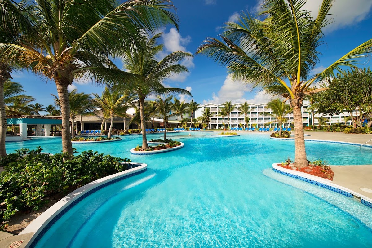 Turquoise lagoon-style pool with palm trees and islands, white resort buildings and blue loungers visible beyond under clear sky.
