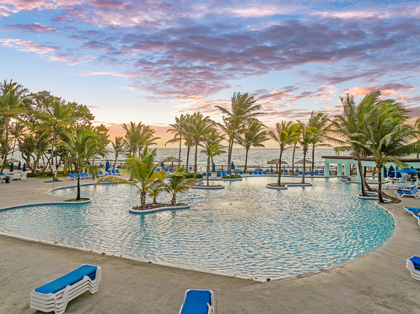 Curved lagoon pool with palm trees dotted throughout at Coconut Bay Beach Resort & Spa, with beach and ocean beyond under a pink-streaked dawn sky.