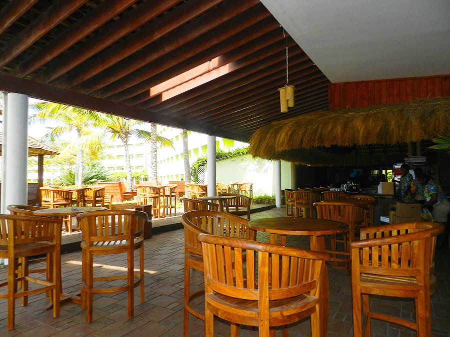 Covered outdoor dining area with wooden chairs and tables, palm trees visible beyond, thatched roof bar structure on right.