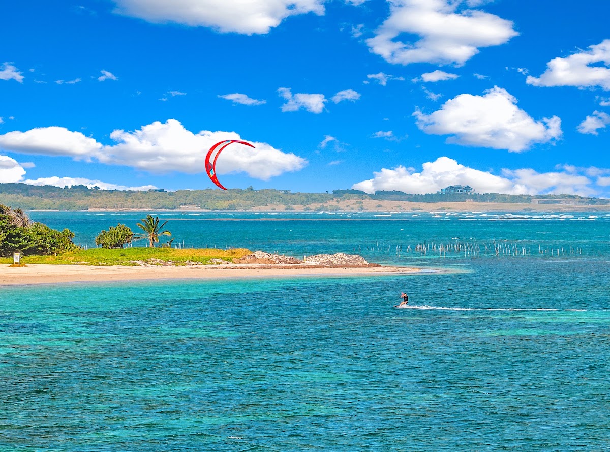 Kiteboarder with red kite in turquoise water off Coconut Bay Beach Resort & Spa, with sandy beach and green hillside beyond.