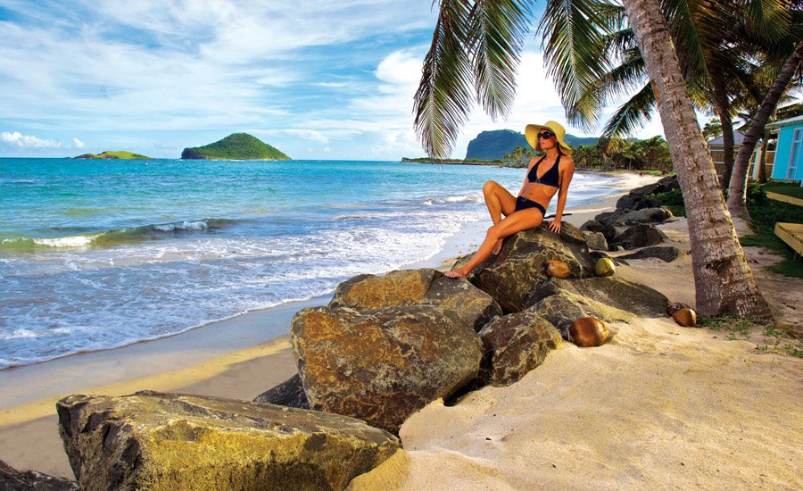 Woman in yellow hat sitting on rocks along sandy beach with palm trees, turquoise water, and green islands visible at Coconut Bay Beach Resort & Spa.