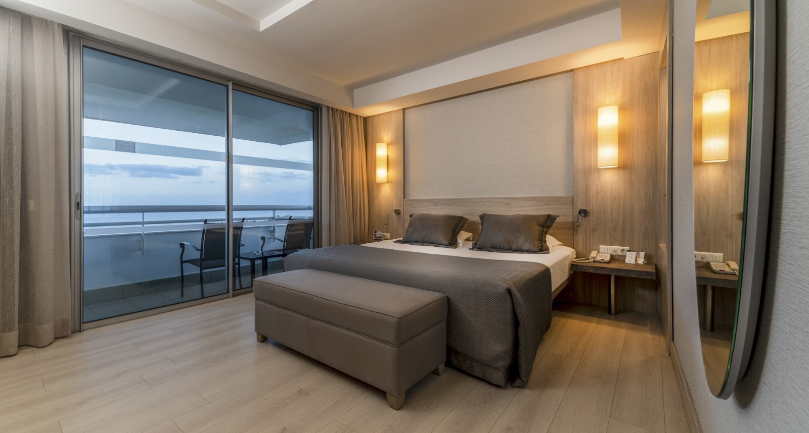 Modern resort bedroom with ocean view balcony, king bed with gray bedding, wooden flooring, and illuminated headboard wall.