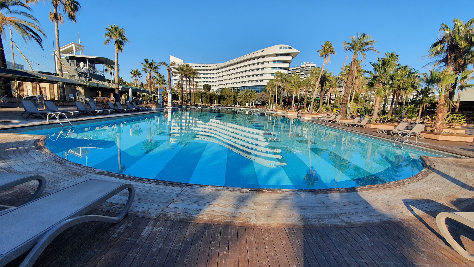 Large curved pool with blue water surrounded by wooden deck, lounge chairs, palm trees, and a multi-story resort building under clear sky.