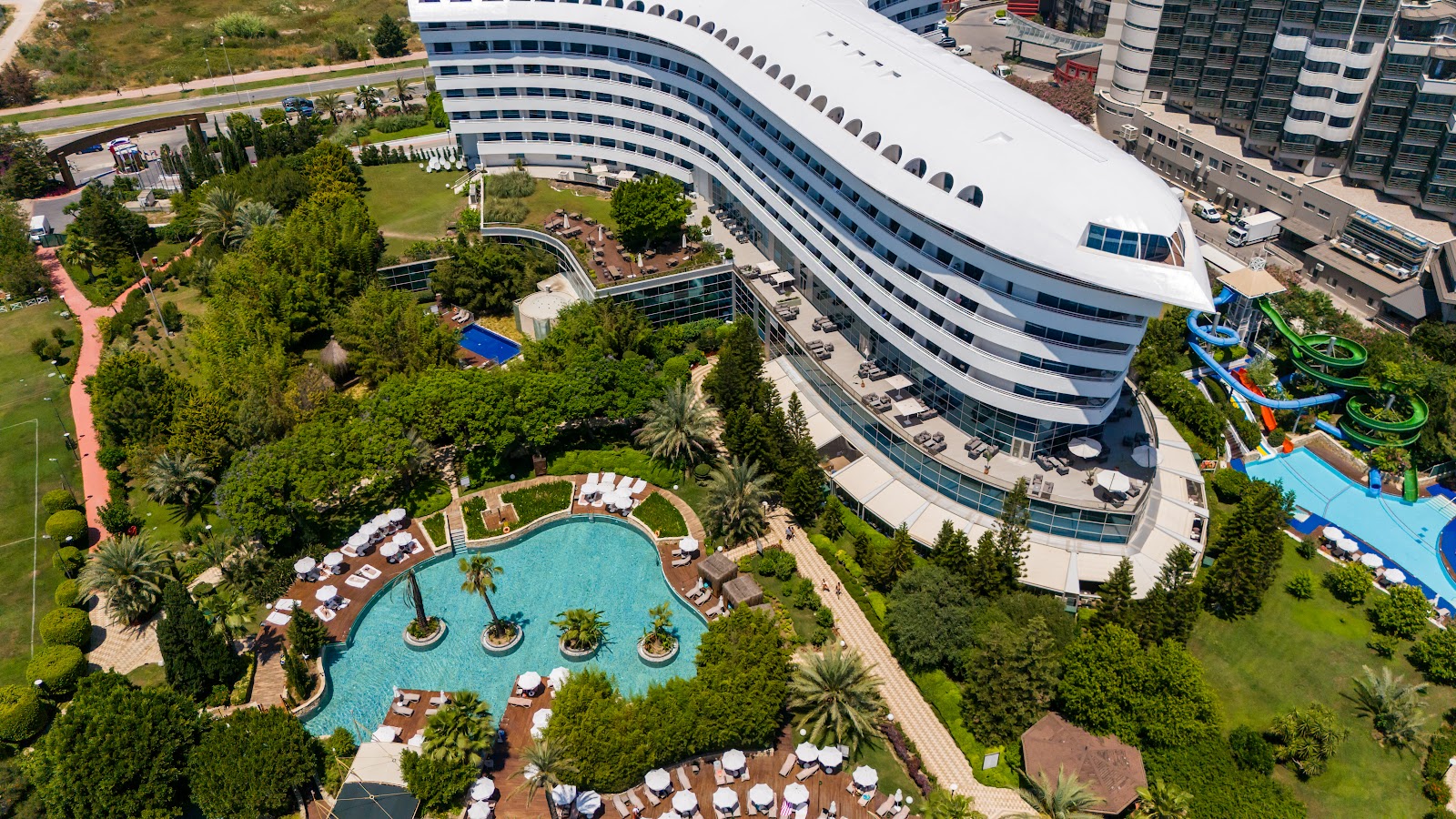 Aerial view of Concorde De Luxe Resort's curved white building, lagoon pool with palm trees, water slides, and manicured grounds surrounded by landscaping.