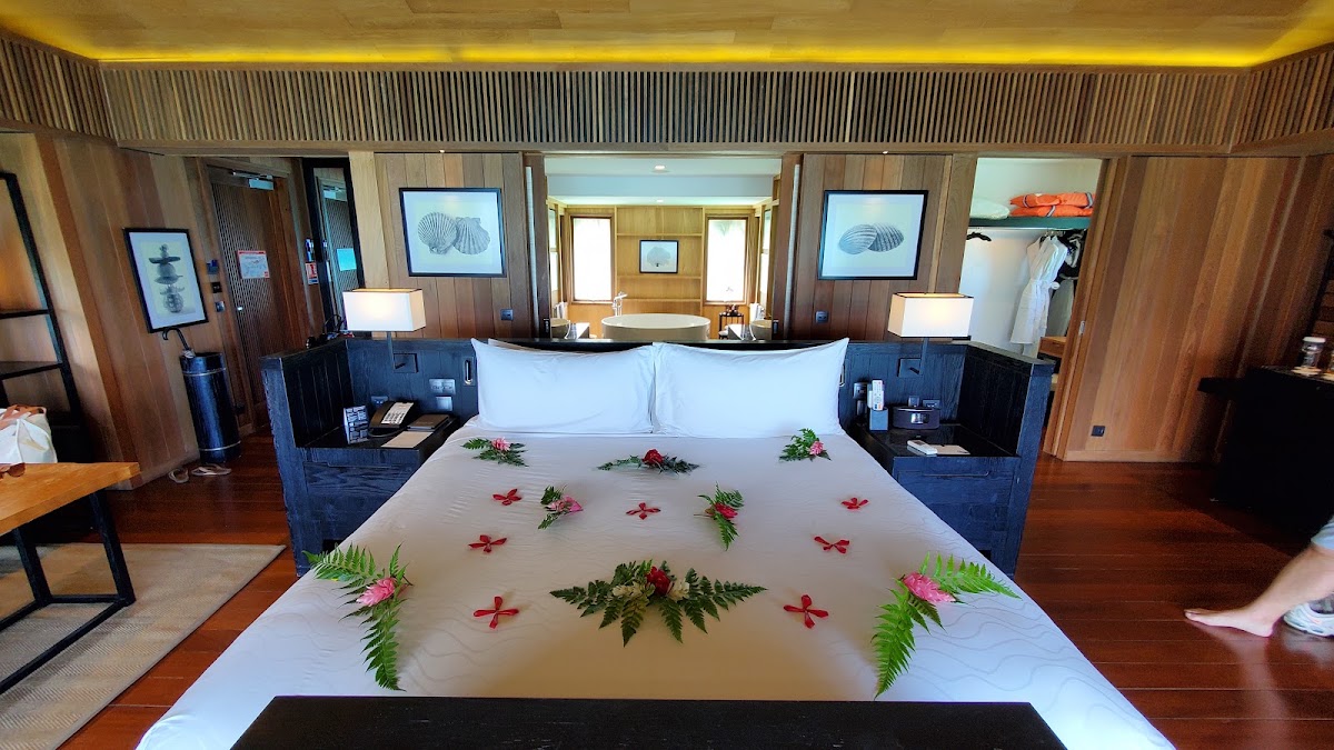 Conrad Bora Bora Nui overwater villa bedroom with king bed decorated with red flowers and green ferns, blue nightstands, wood paneling, and ocean views through windows.