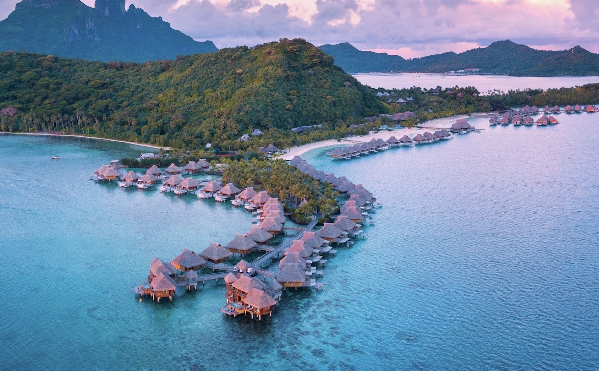 Overwater bungalows with thatched roofs line turquoise lagoon waters at Conrad Bora Bora Nui, with lush green mountains and a pink sunset sky beyond.