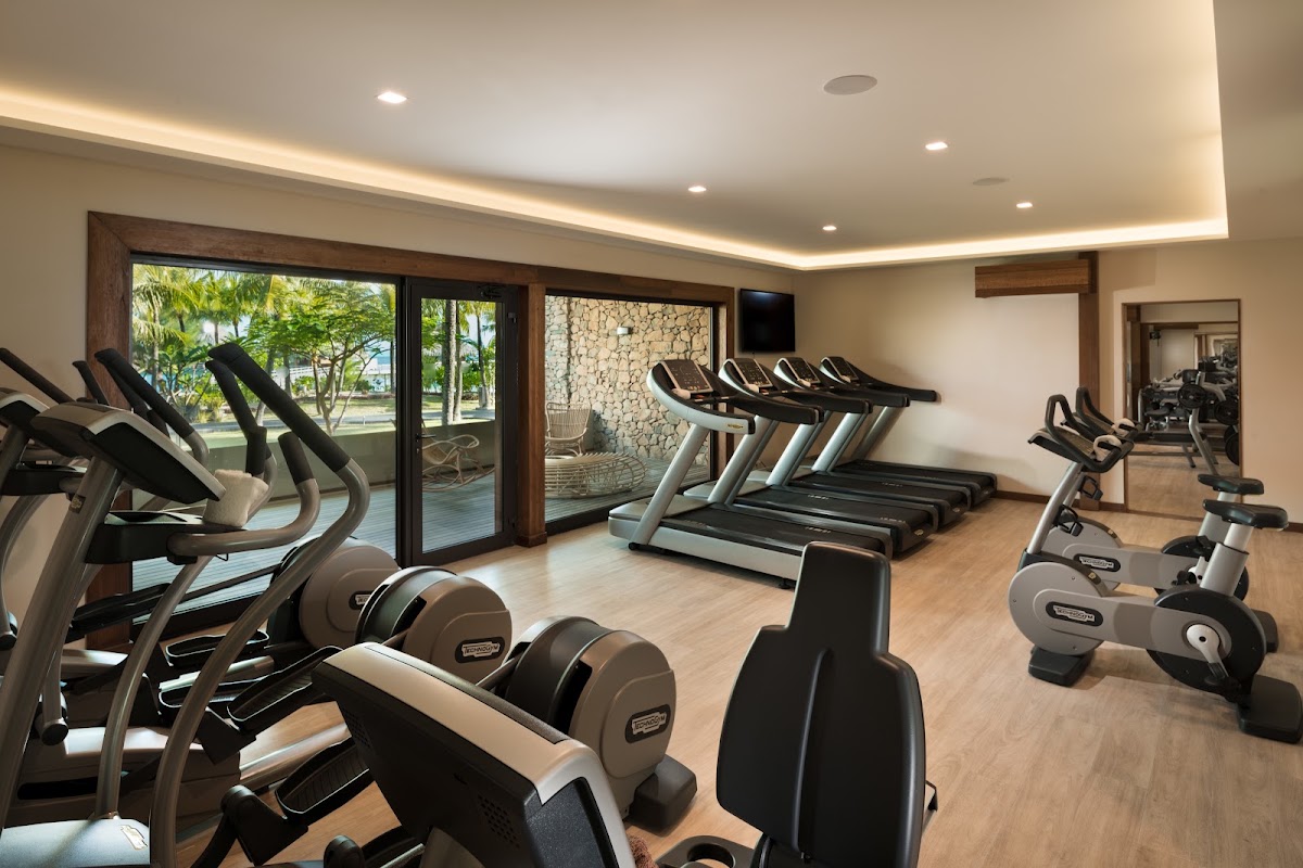 Resort fitness center with treadmills, stationary bikes, and dumbbells; glass doors open to tropical grounds with palm trees.
