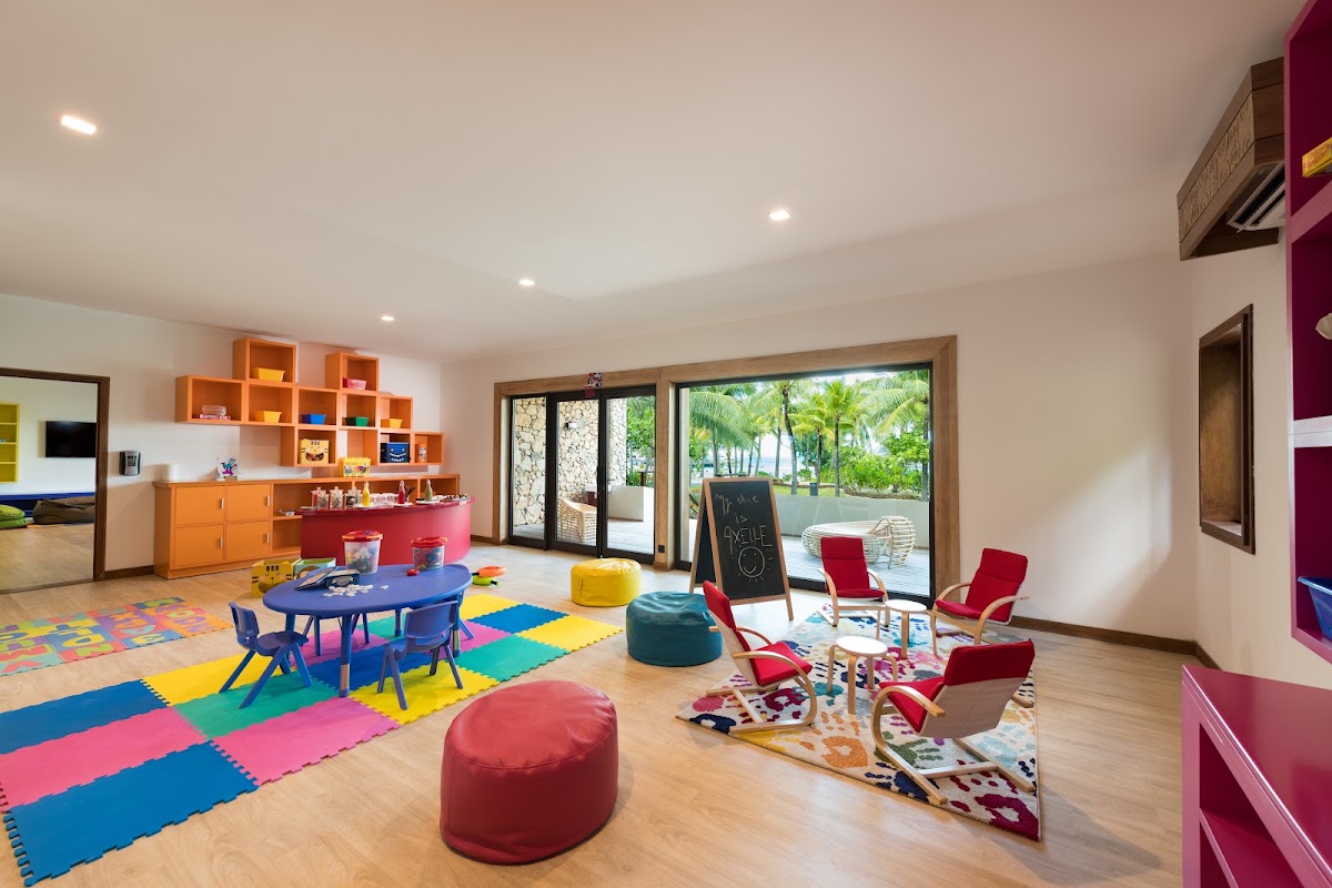 Colorful children's playroom with blue table, foam floor mats, orange storage shelves, and glass doors opening to tropical gardens.