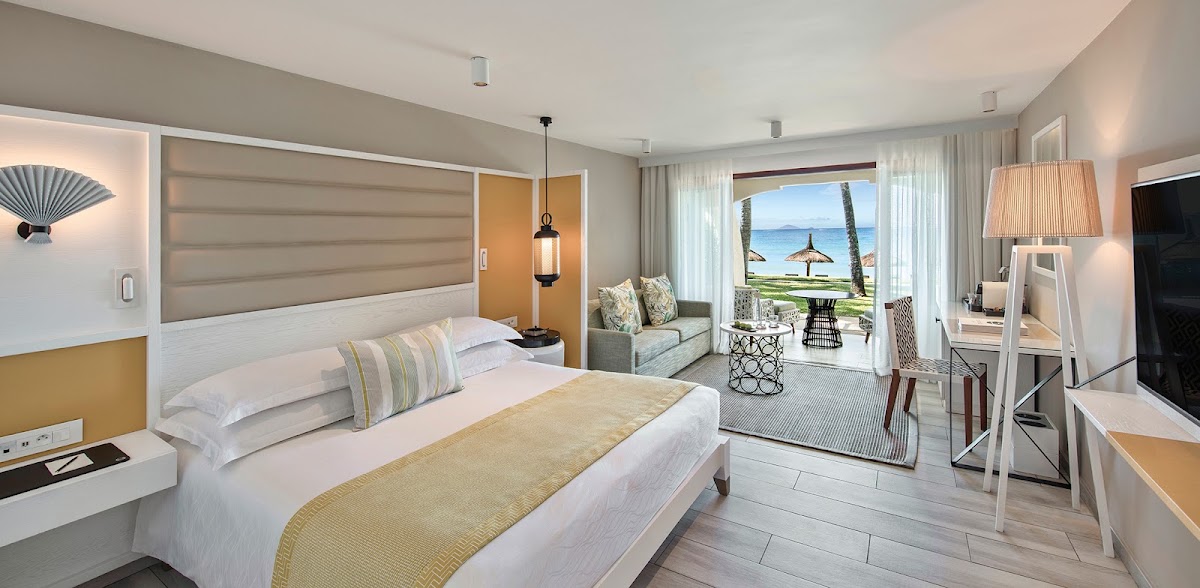 Resort bedroom with ocean view from balcony, featuring beige upholstered headboard, white bedding, and seating area overlooking beach with umbrellas.